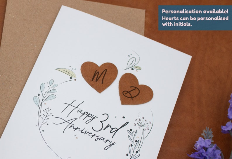 K&ouml;nnte beinhalten: Eine wei&szlig;e Karte mit einem floralen Design und dem Text "Happy 3rd Anniversary". Zwei braune herzf&ouml;rmige Papierschnitte mit den Initialen "M" und "D" sind an der Karte befestigt.