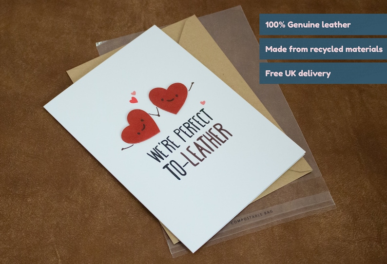 May include: A white card with the text "WE'RE PERFECT TO-LEATHER" and two red hearts with faces. The card is on a brown surface and has a clear plastic bag with the text "COMPOSTABLE BAG" on it. The card is made from recycled materials and is 100% genuine leather.