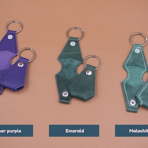 May include: Three sets of keychains in biker purple, emerald, and malachite colors. Each set includes a larger geometric-shaped leather piece with a metal ring and a smaller matching leather piece with a metal ring, both secured by silver snaps.