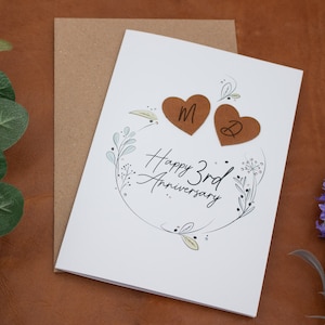K&ouml;nnte beinhalten: Eine wei&szlig;e Karte mit einem floralen Design und dem Text "Happy 3rd Anniversary". Zwei braune Lederherzen mit den Initialen "M" und "D" sind an der Karte befestigt.