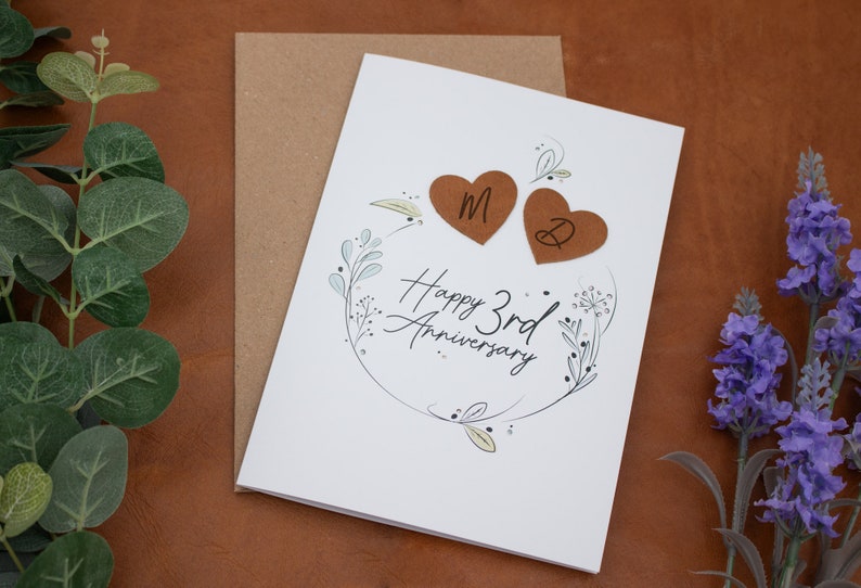 K&ouml;nnte beinhalten: Eine wei&szlig;e Karte mit einem floralen Design und dem Text "Happy 3rd Anniversary". Zwei braune Lederherzen mit den Initialen "M" und "D" sind an der Karte befestigt.