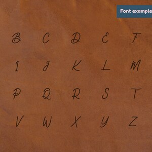 K&ouml;nnte beinhalten: Ein handgeschriebenes Alphabet in schwarzer Tinte auf braunem Lederhintergrund. Die Buchstaben sind in einem l&auml;ssigen, kursiven Stil geschrieben.