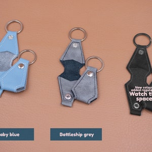 May include: Three leather keychains in baby blue, battleship grey, and black. Each features a metal ring and a snap-button closure. The black keychain displays the text: New colours added regularly... Watch this space! The items are arranged in a row.