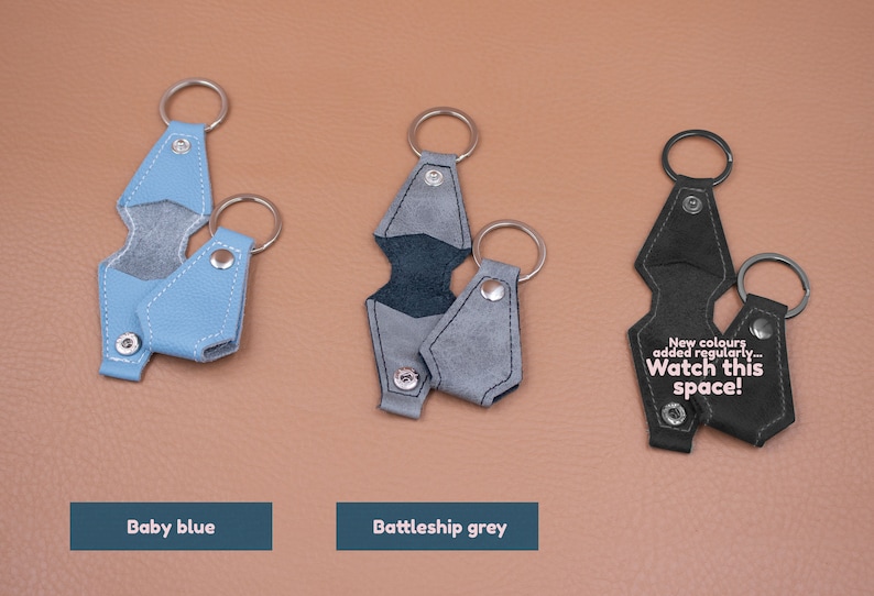 May include: Three leather keychains in baby blue, battleship grey, and black. Each features a metal ring and a snap-button closure. The black keychain displays the text: New colours added regularly... Watch this space! The items are arranged in a row.