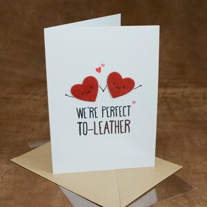 May include: A white card with two red felt hearts holding hands. The hearts have smiley faces. The text on the card reads "We're perfect to-leather".