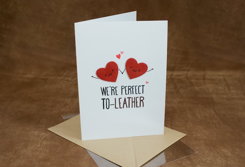 May include: A white card with two red felt hearts holding hands. The hearts have smiley faces. The text on the card reads "We're perfect to-leather".