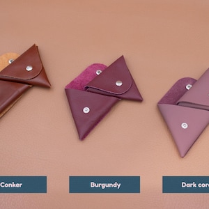 May include: Three leather triangle-shaped coin purses in shades of brown, burgundy, and dark coral. Each purse has a snap closure and a contrasting interior lining. The purses are displayed on a neutral background.