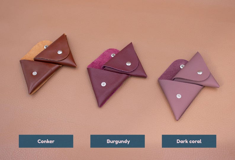 May include: Three leather triangle-shaped coin purses in shades of brown, burgundy, and dark coral. Each purse has a snap closure and a contrasting interior lining. The purses are displayed on a neutral background.