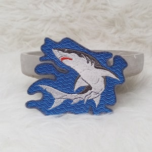 Könnte beinhalten: Ein gestickter Aufnäher mit einem weißen Hai mit rotem Maul und schwarzen Augen, der aus blauem Wasser mit weißen Wellen springt. Der Aufnäher befindet sich auf einer grauen Oberfläche.