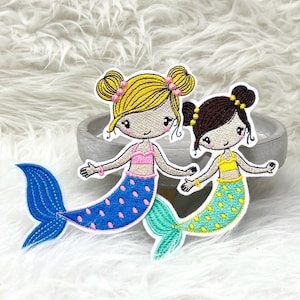 Könnte beinhalten: Zwei gestickte Bügelbilder von Meerjungfrauen mit gelben Haaren, rosa-blauen Tupfen-Schwänzen und gelb-grünen Tupfen-Schwänzen.