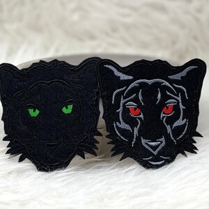 Könnte beinhalten: Zwei schwarze gestickte Patches mit Panthergesichtern und unterschiedlichfarbigen Augen. Ein Panther hat grüne Augen und der andere rote Augen.