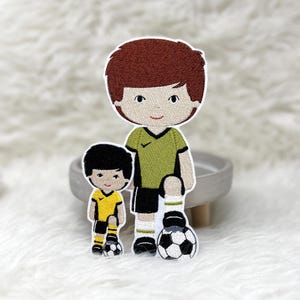 Könnte beinhalten: Zwei aufgestickte Aufnäher von Fußballspielern. Der größere Aufnäher zeigt einen Spieler in einem grünen Trikot, schwarzen Shorts und weißen Socken, der einen Fußball hält. Der kleinere Aufnäher zeigt einen Spieler in einem gelben Trikot und schwarzen Shorts, ebenfalls mit einem Fußball.