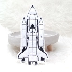 Könnte beinhalten: Ein weißer gestickter Aufnäher einer Raumfähre mit schwarzen Umrissen. Die Fähre hat zwei Seitenbooster und einen Hauptmotor.
