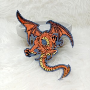 Könnte beinhalten: Orangefarbener gestickter Drache mit ausgebreiteten Flügeln auf blauem Hintergrund. Der Drache befindet sich in einer zusammengerollten Position mit ausgestreckten Krallen.