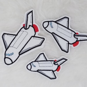 Könnte beinhalten: Drei weiße gestickte Patches von Raumschiffen mit roten und blauen Details. Die Patches befinden sich auf einer weißen, flauschigen Oberfläche.