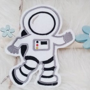 Könnte beinhalten: Ein weißer Astronauten-Applik mit schwarzen und grauen Details, der mit einem erhobenen Bein geht. Der Astronaut hat einen runden Helm mit einem schwarzen Visier. Der Applik befindet sich auf einem weißen, flauschigen Hintergrund.