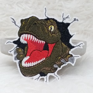 Könnte beinhalten: Gestickter Aufnäher mit einem grünen und braunen Dinosaurier, der durch ein schwarzes Loch bricht. Der Dinosaurier hat einen roten Mund mit weißen Zähnen und gelben Augen. Der Aufnäher befindet sich auf weißem Hintergrund.