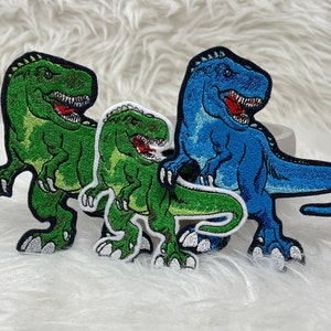 Könnte beinhalten: Drei gestickte Bügelbilder mit Cartoon-Dinosauriern. Die Dinosaurier sind grün, blau und grün. Die Patches sind perfekt, um Kleidung, Taschen oder anderen Stoffartikeln eine lustige und fantasievolle Note zu verleihen.