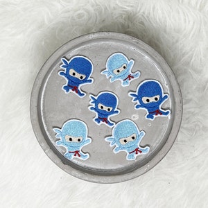 Könnte beinhalten: Sechs blaue und weiße gestickte Bügelbilder von Ninjas in verschiedenen Posen. Die Patches befinden sich auf einer grauen Oberfläche.