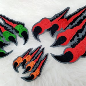 Könnte beinhalten: Vier gestickte Bügelbilder mit verschiedenen farbigen Krallen. Die Krallen sind rot, grün, orange und schwarz. Die Patches befinden sich auf einer weißen, flauschigen Oberfläche.