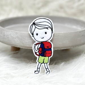 Könnte beinhalten: Gestickter Aufnäher eines Cartoon-Jungen mit rotem Rucksack, grünen Shorts und weißem Hemd. Der Junge lächelt und hält ein Buch.