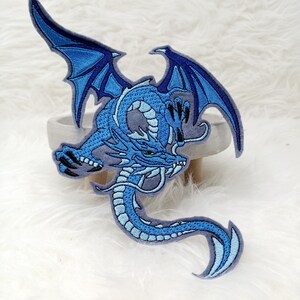 Könnte beinhalten: Blauer gestickter Bügelaufkleber mit einem Drachen mit ausgebreiteten Flügeln. Der Drache ist in einer zusammengerollten Position mit dem Kopf nach vorne und dem Maul offen.