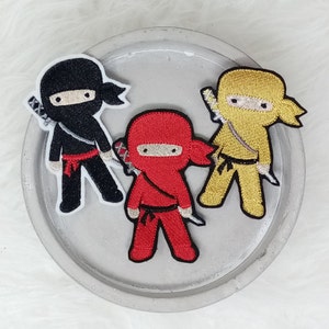 Könnte beinhalten: Drei gestickte Bügelbilder mit Cartoon-Ninjas in Schwarz, Rot und Gelb. Jeder Ninja trägt eine Maske und hält ein Schwert.