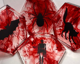 Puede incluir: Conjunto de cuatro posavasos hexagonales con un diseño marmoleado rojo y blanco. Cada posavasos presenta una silueta negra de una criatura de Halloween: una araña, un murciélago, un esqueleto y un gato.
