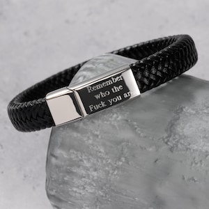 Könnte beinhalten: Schwarzes geflochtenes Lederarmband mit silbernem Verschluss. Der Verschluss ist mit den Worten "Remember who the Fuck you are" graviert. Das Armband liegt auf einer grauen Steinoberfläche.