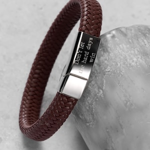 Könnte beinhalten: Ein geflochtenes Armband aus braunem Leder mit einem silbernen Verschluss. Auf dem Verschluss steht "Don't let the hard days win". Das Armband liegt auf einer hellgrauen Oberfläche.