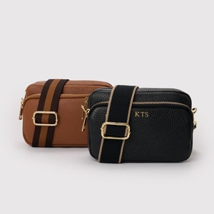May include: Two small leather crossbody bags with adjustable straps. The bag on the left is brown with a black and brown striped strap. The bag on the right is black with a black and beige striped strap. The black bag has the initials "KTS" embossed on the front.