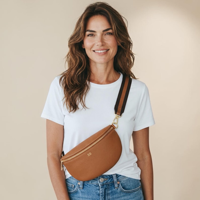 K&ouml;nnte beinhalten: Eine hellbraune Bauchtasche aus Leder mit goldenem Rei&szlig;verschluss und den eingepr&auml;gten Initialen SB auf der Vorderseite. Die Tasche hat einen breiten, verstellbaren Stoffgurt mit braun-schwarzen Streifen und goldfarbenen Metallbeschl&auml;gen.