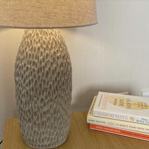 May include: A beige ceramic table lamp with a textured base and a neutral linen lampshade. Several books are stacked on a wooden surface next to the lamp.
