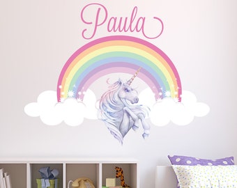 May include: A white unicorn with a rainbow and clouds wall decal. The rainbow is in pastel colors and the unicorn is standing on its hind legs. The name "Paula" is written in pink cursive above the rainbow.