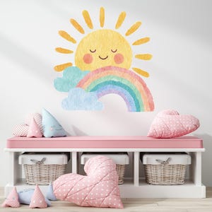 May include: A colourful wall decal of a smiling sun, rainbow, and clouds. A pink bench with white storage baskets and heart-shaped pillows in pink and white dots. Decorative pillows in various shapes and colours.