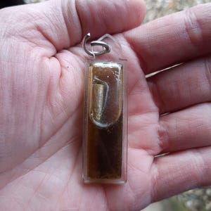 Puede incluir: Un colgante rectangular con un interior marrón y dorado, encerrado en plástico transparente. El colgante tiene un lazo de metal plateado en la parte superior para sujetarlo a una cadena o llavero. El colgante se sostiene en una mano.