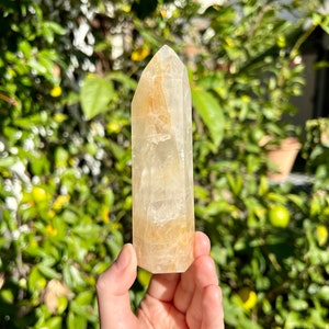 Puede incluir: Una punta de cristal de cuarzo transparente con un tono dorado. El cristal mide aproximadamente 10 cm de alto y tiene una superficie lisa y pulida.