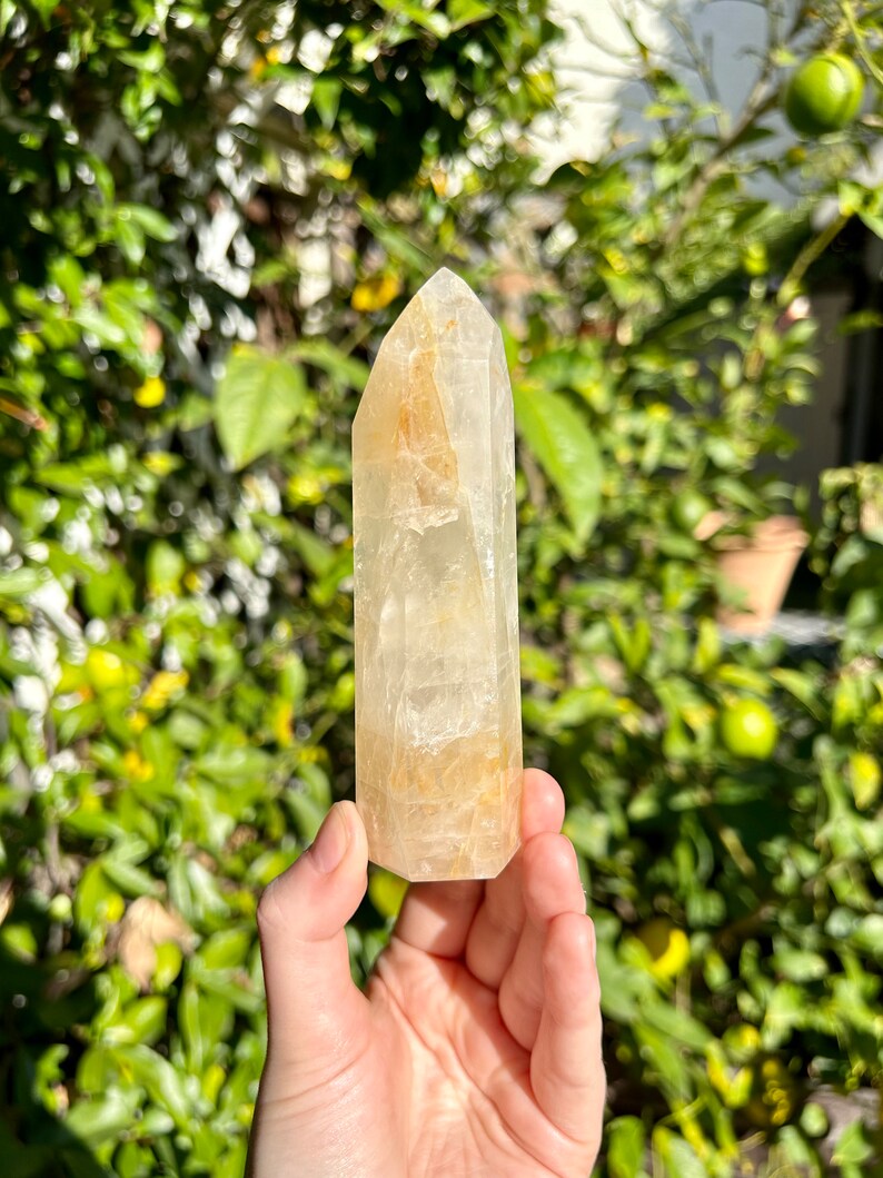 Puede incluir: Una punta de cristal de cuarzo transparente con un tono dorado. El cristal mide aproximadamente 10 cm de alto y tiene una superficie lisa y pulida.