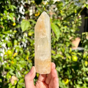 Puede incluir: Una punta de cristal de cuarzo transparente con un tono dorado. El cristal mide aproximadamente 13 cm de alto y tiene una superficie lisa y pulida.