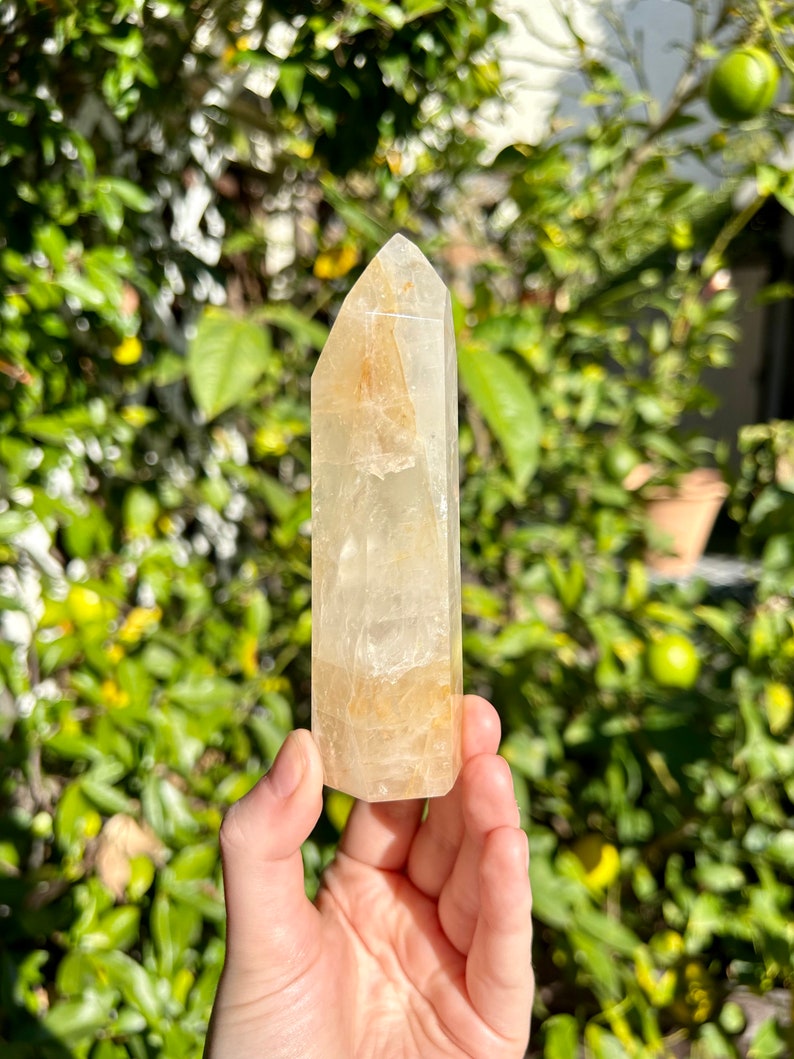 Puede incluir: Una punta de cristal de cuarzo transparente con un tono dorado. El cristal mide aproximadamente 13 cm de alto y tiene una superficie lisa y pulida.