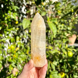 Puede incluir: Una punta de cristal transparente y facetada con un tono amarillo-naranja. El cristal mide aproximadamente 12,5 cm de alto y tiene una punta puntiaguda. Se sostiene en una mano contra un fondo borroso de follaje verde.