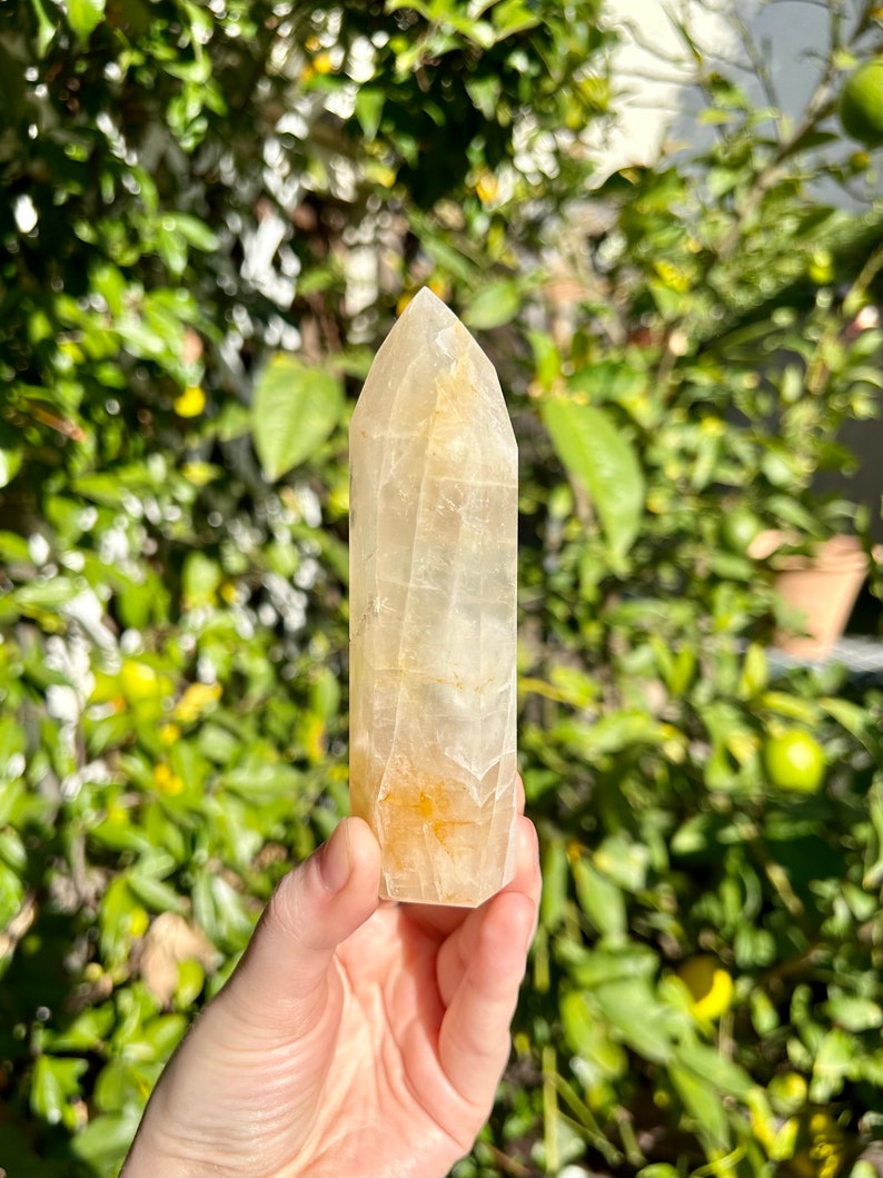 Puede incluir: Una punta de cristal transparente y facetada con un tono amarillo-naranja. El cristal mide aproximadamente 12,5 cm de alto y tiene una punta puntiaguda. Se sostiene en una mano contra un fondo borroso de follaje verde.
