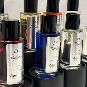 May include: A collection of eight small glass bottles with black caps and silver labels. The labels feature the text "B.C. Parfums" and a skull graphic. The bottles are filled with different colored liquids.