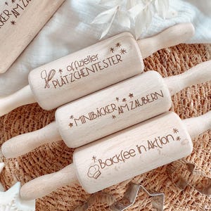 Könnte beinhalten: Holz-Nudelrollen mit graviertem Text und Sternen. Eine Rolle trägt die Aufschrift "offizieller PLATZCHENTESTER", eine andere "MINIBÄCKER AM ZAUBERN" und eine dritte "Backfee in Aktion". Die Nudelrollen werden mit sternförmigen Ausstechern präsentiert.