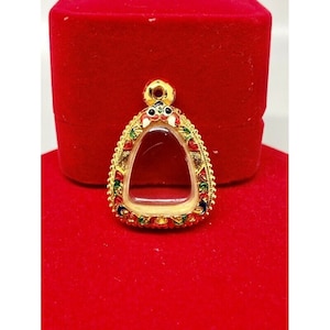 Könnte beinhalten: Ein goldfarbener Anhänger mit einem transparenten Mittelteil, umgeben von aufwendigen bunten Designs. Der Anhänger hat eine Tropfenform und ist vor rotem Hintergrund platziert, was auf Schmuck oder Accessoires hindeutet.