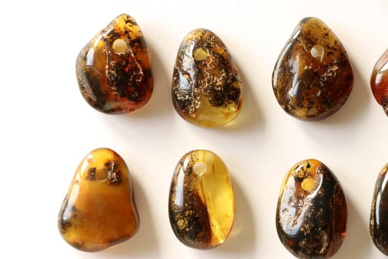 May include: A collection of eight amber gemstone pendants with a hole drilled through the top for stringing. The pendants are various shades of amber, from light honey to dark brown, and some have inclusions of black or white.