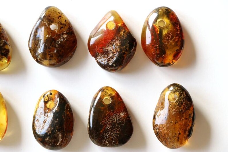 May include: Ten amber gemstone pendants with a hole drilled through the top. The pendants are various shades of brown, yellow, and orange. Some of the pendants have inclusions of insects or other organic matter.