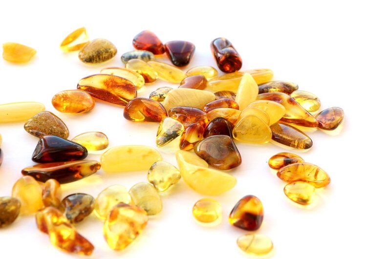 May include: A collection of amber stones in various shades of yellow, orange, and brown. The stones are smooth and polished, and they are scattered on a white background.