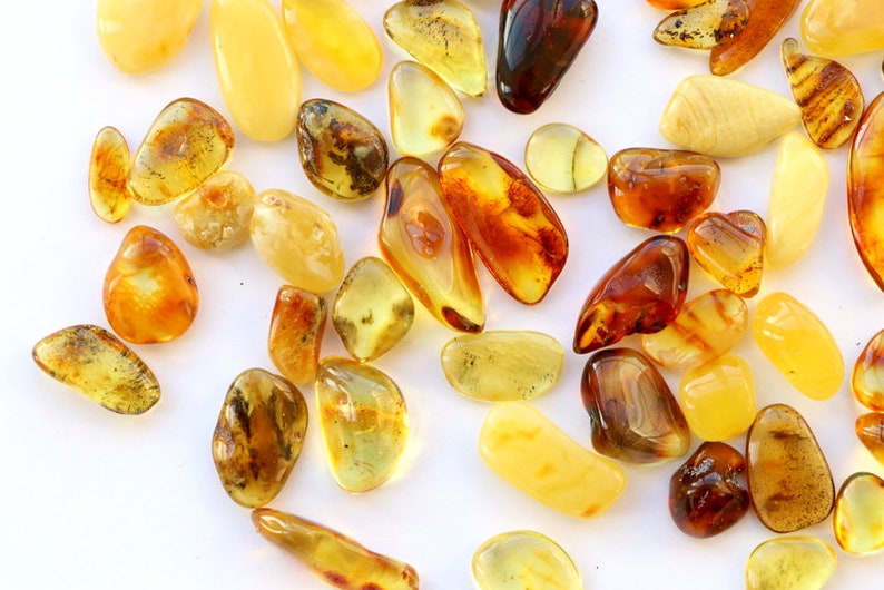 May include: A collection of amber stones in various shades of yellow, orange, and brown. The stones are smooth and polished, with some showing inclusions of insects or plant matter.