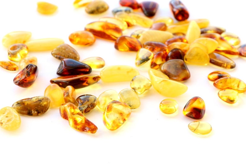 May include: A collection of amber stones in various shades of yellow, orange, and brown. The stones are scattered on a white background.